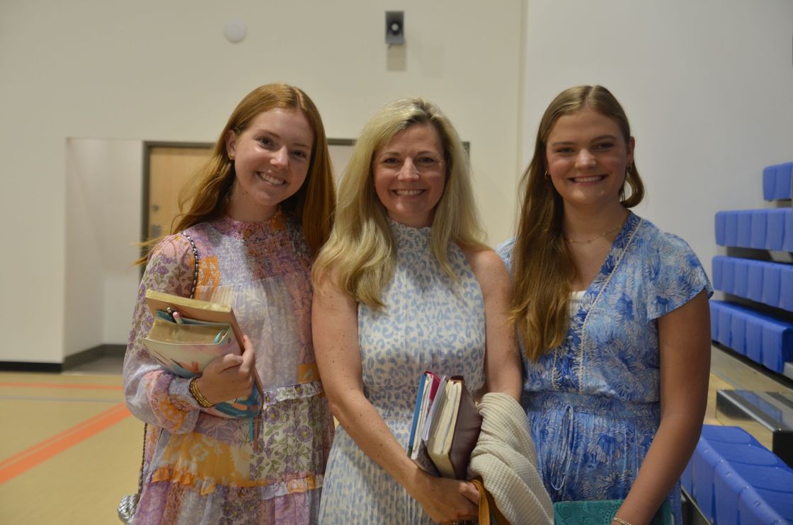 Woman smiles between two young women holding books in a school hallway.