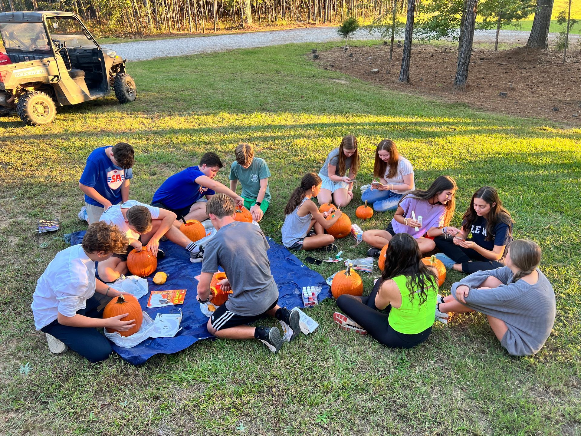 A group of people are sitting in a circle in the grass carving pumpkins.