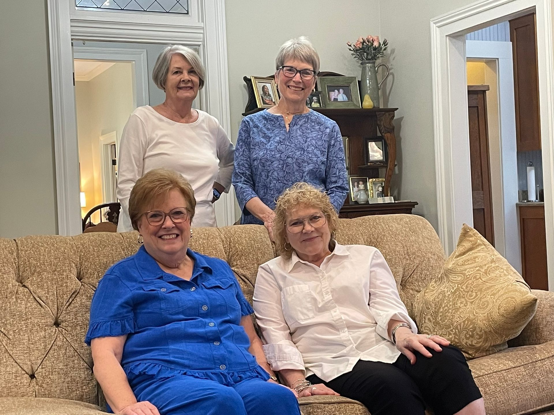 Four women smiling in a living room. Two seated on a couch, two standing behind.