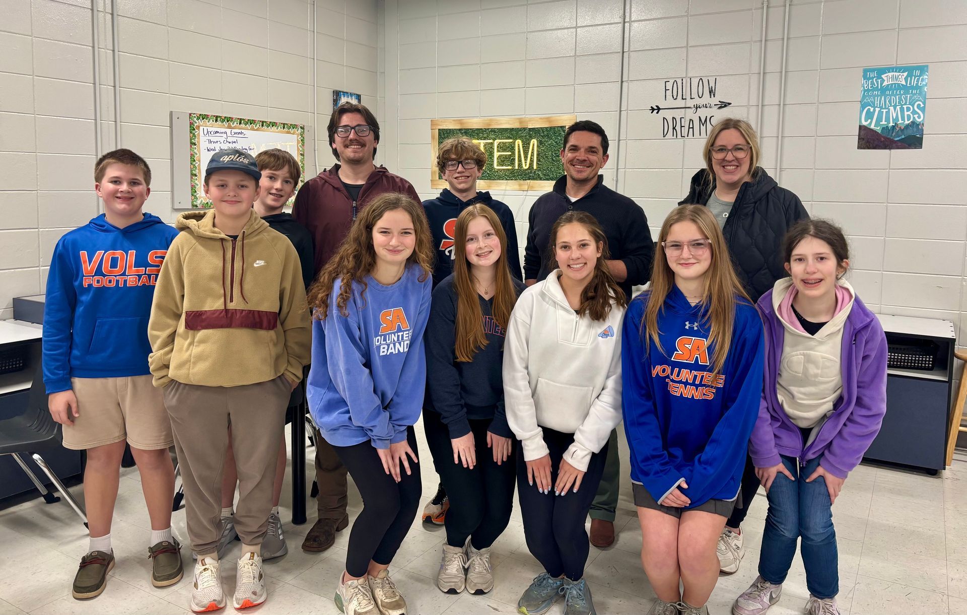 Group of students and adults posing in a classroom; smiles, casual attire, neutral background.