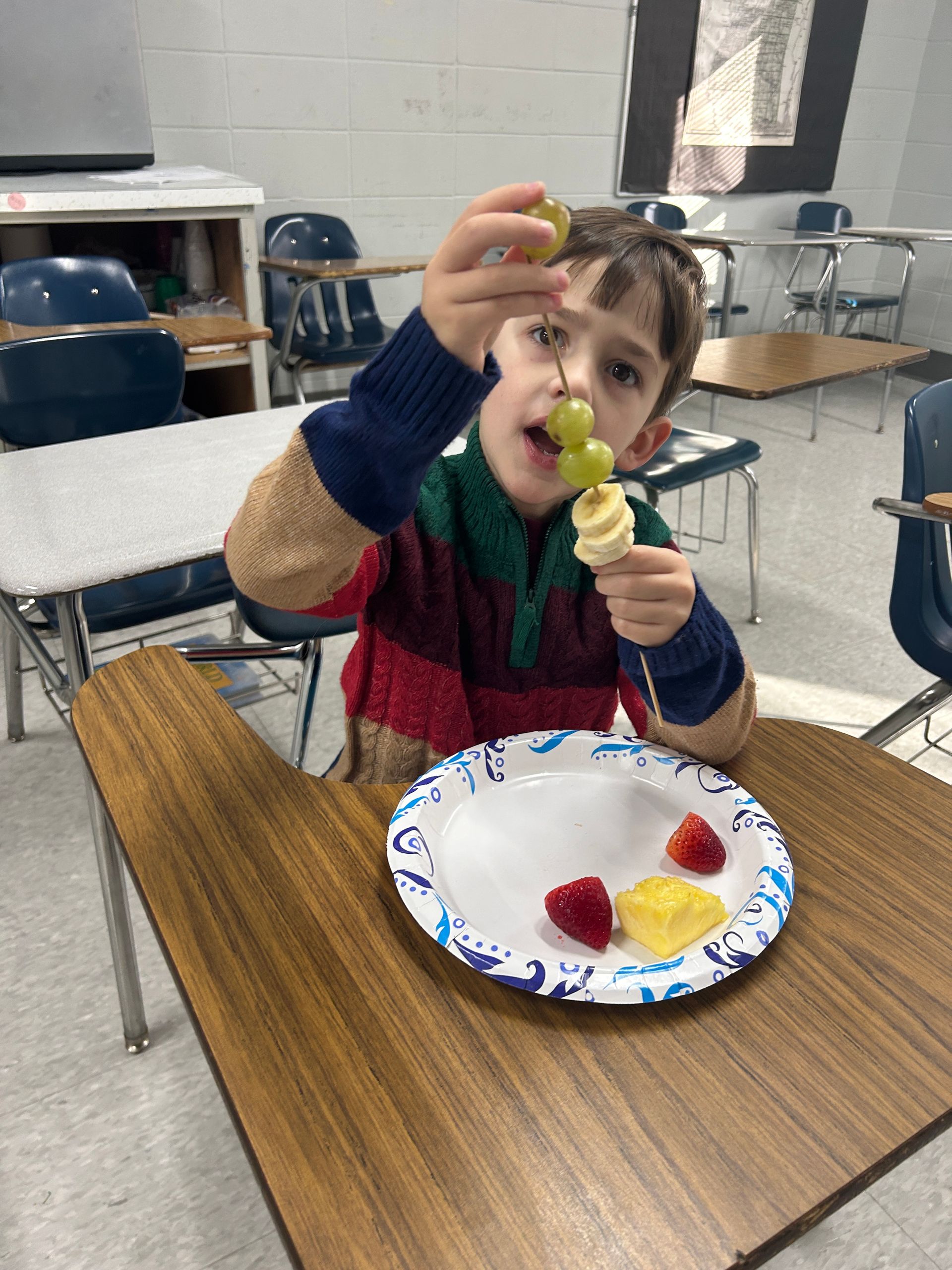 A young boy is sitting at a table eating fruit skewers.