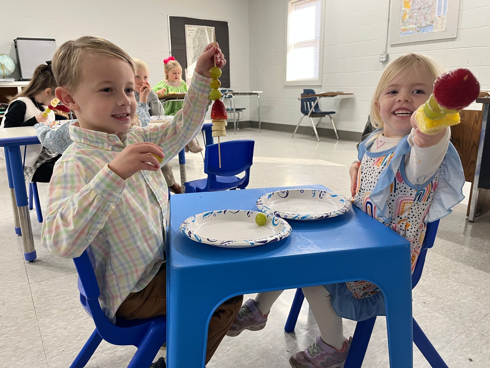 A boy and a girl are sitting at a table holding fruit on skewers.