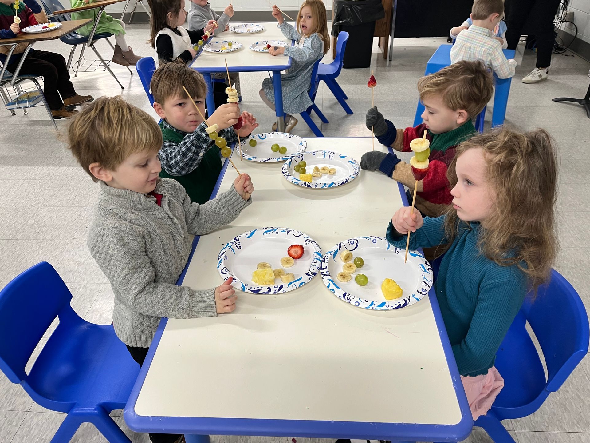A group of children are sitting at a table eating fruit on skewers.