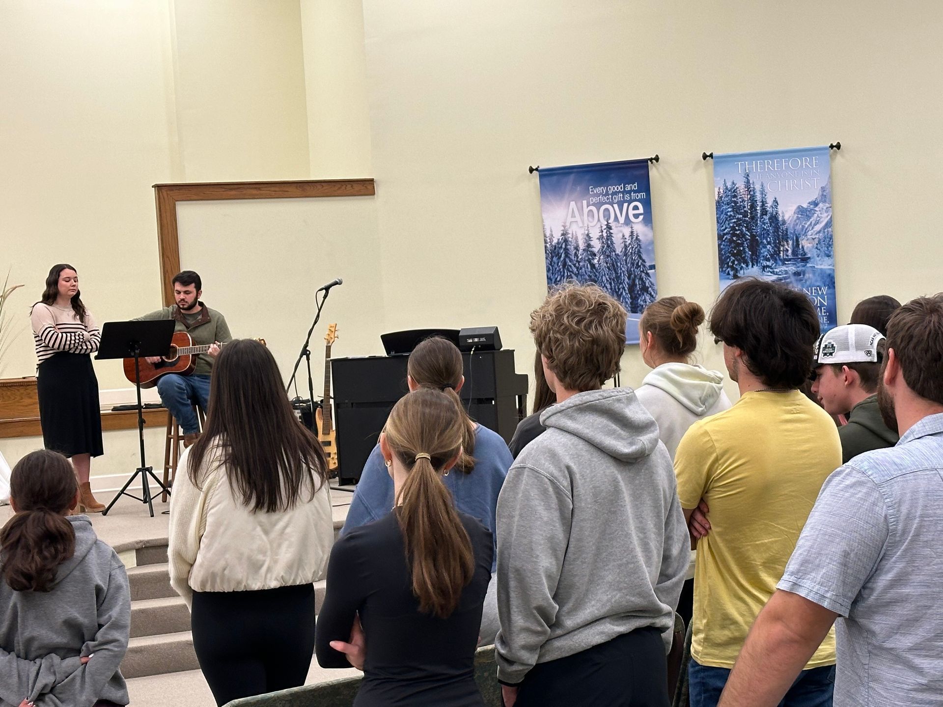 A group of people are standing in a room watching a man play a guitar.