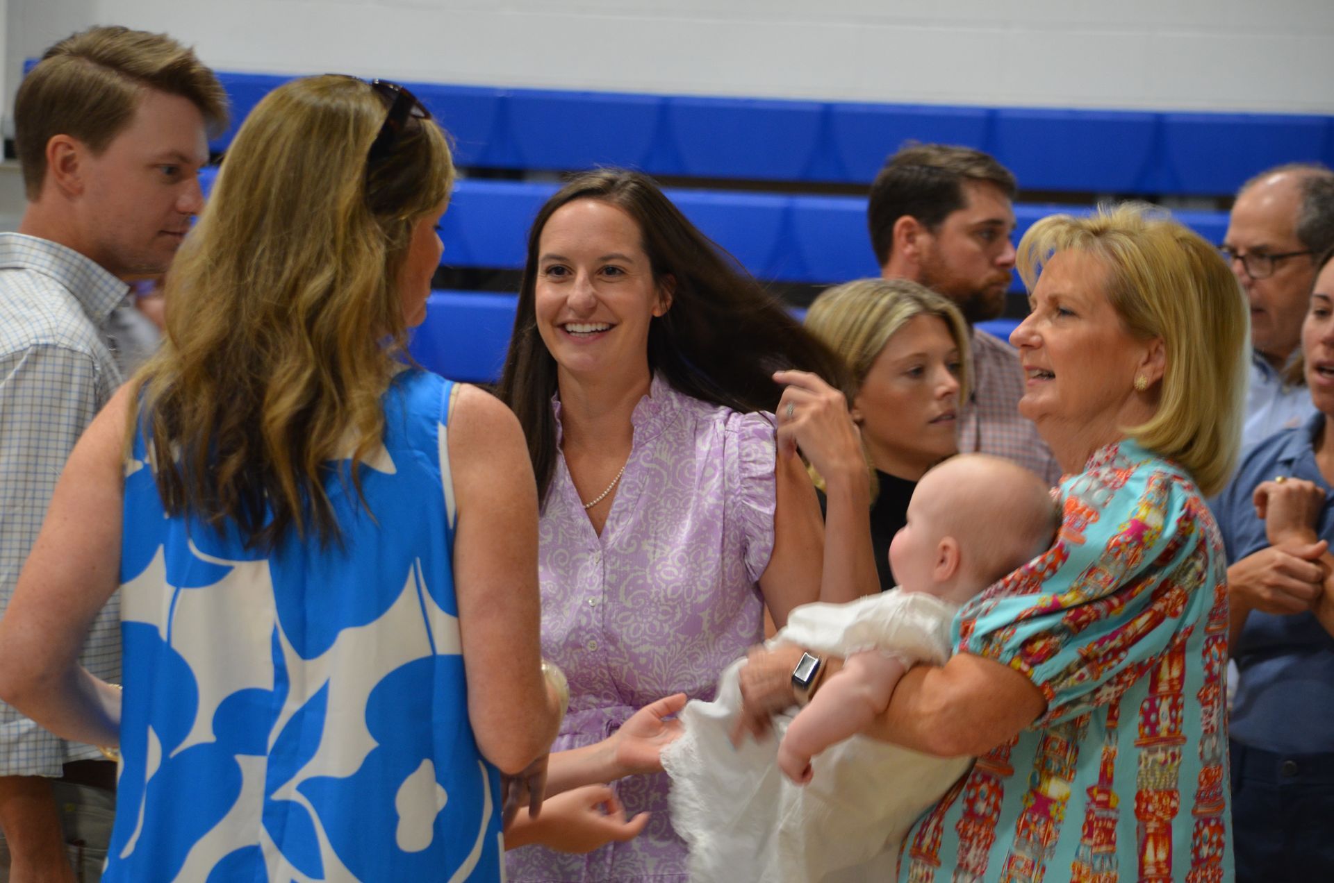 People at an event, one holding a baby in white dress. Blue and floral print dresses, smiles, and a blue wall.