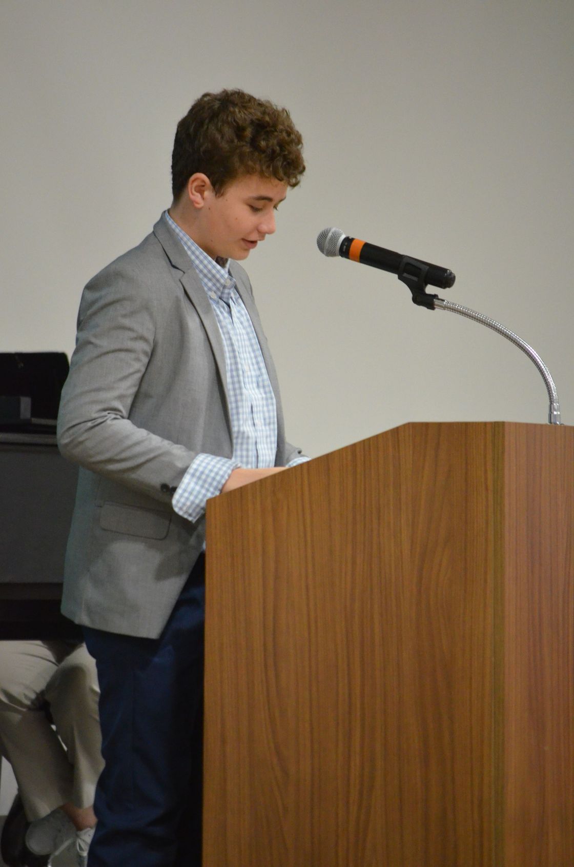 Young person speaking at a podium, wearing a grey blazer and blue shirt.