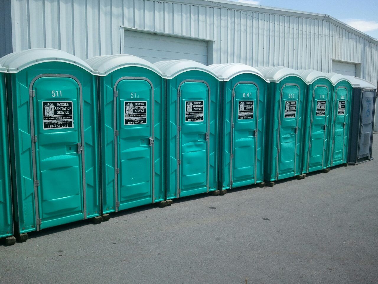 A row of teal portable toilets lined up outdoors against the side of a white industrial building.
