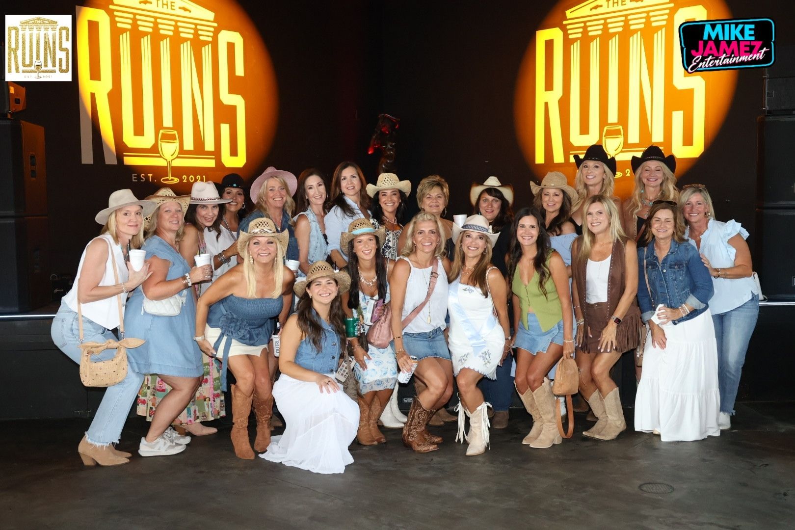 Group of people posing onstage in front of large illuminated “RUMS” signs, many in white and denim outfits