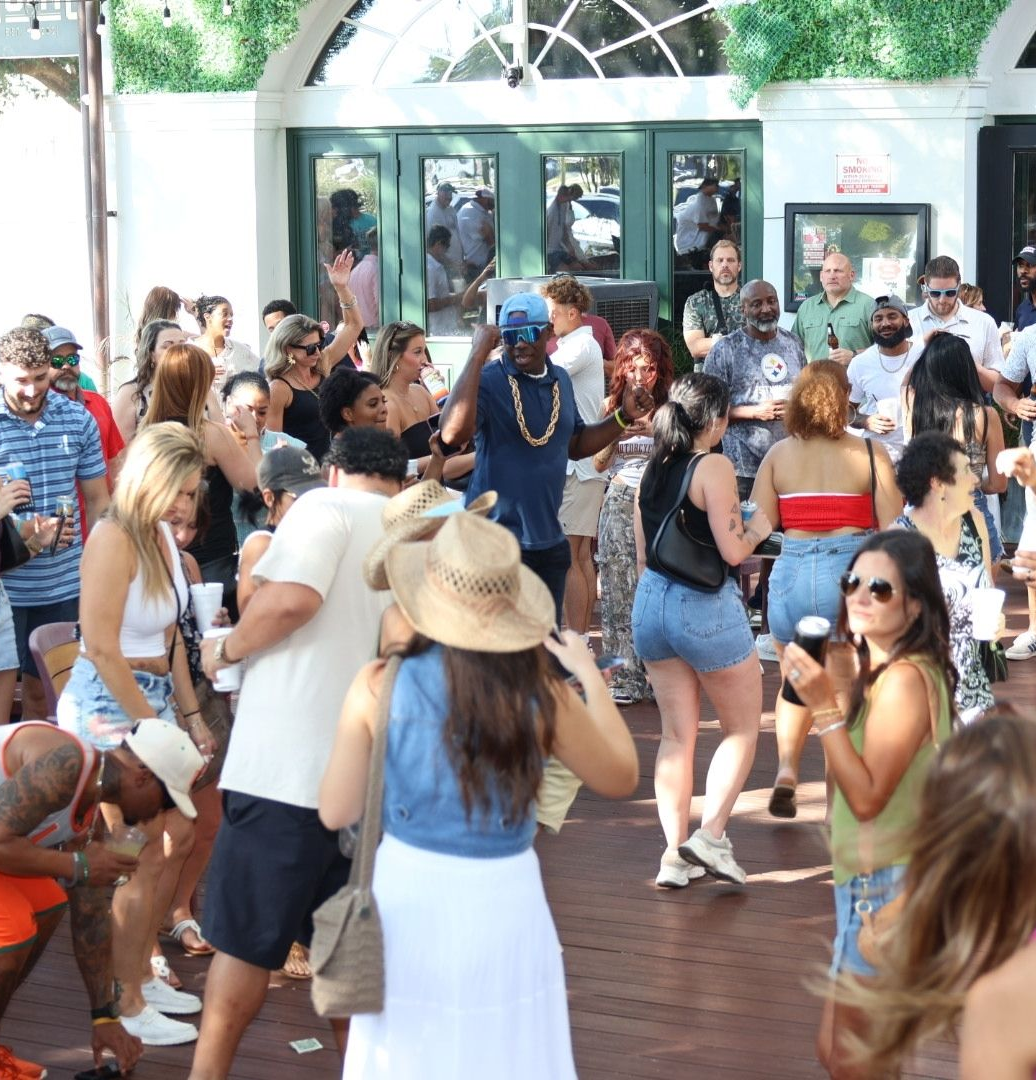Crowded outdoor party with people dancing on a wooden deck outside a glass-front building