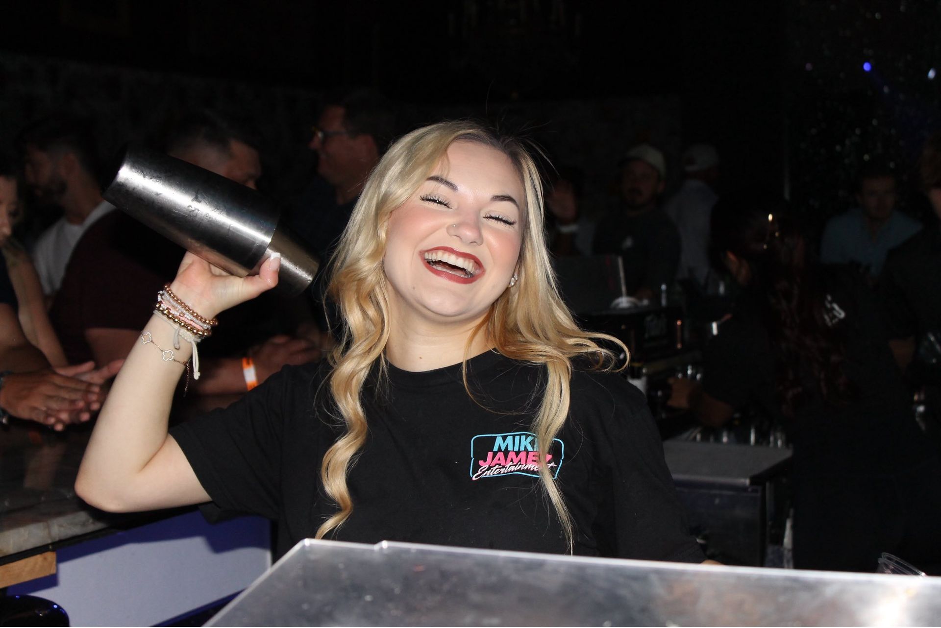 Smiling woman in a black shirt shaking a cocktail shaker at a nightclub booth