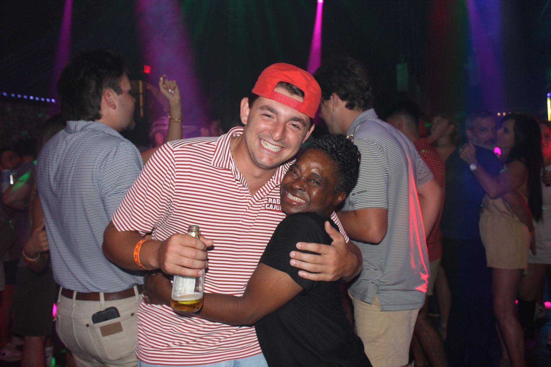 Two people hugging and smiling in a nightclub, one holding a drink and wearing a red cap.