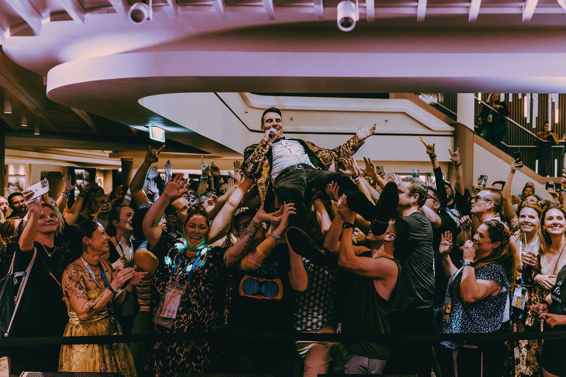 Crowd cheering and lifting a person in a bright indoor venue with a staircase and warm lighting