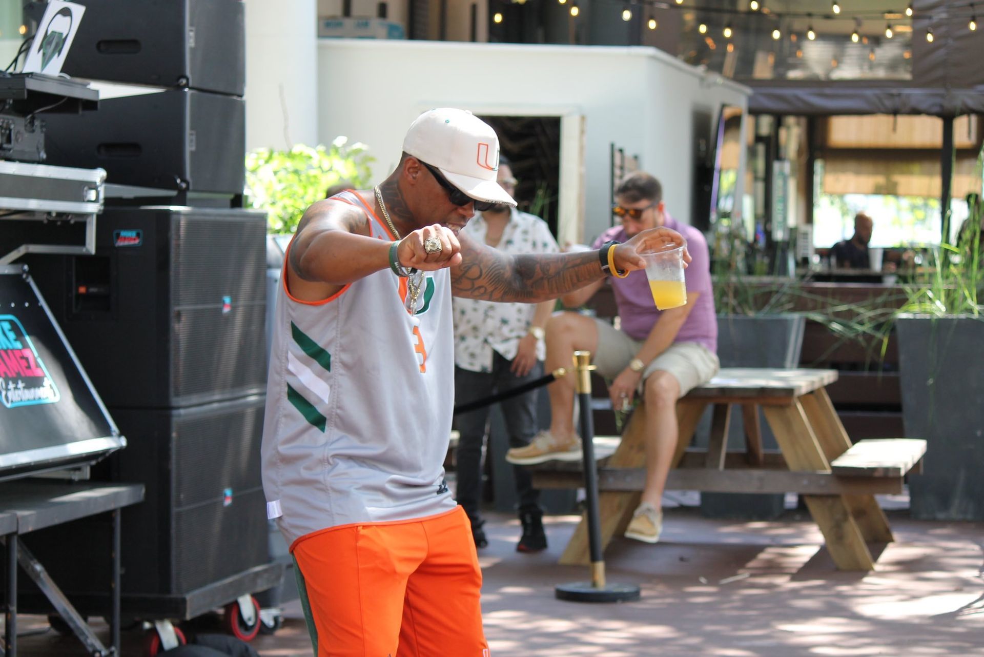 Man pointing toward the camera in a bright outdoor patio area, with a seated person behind him.