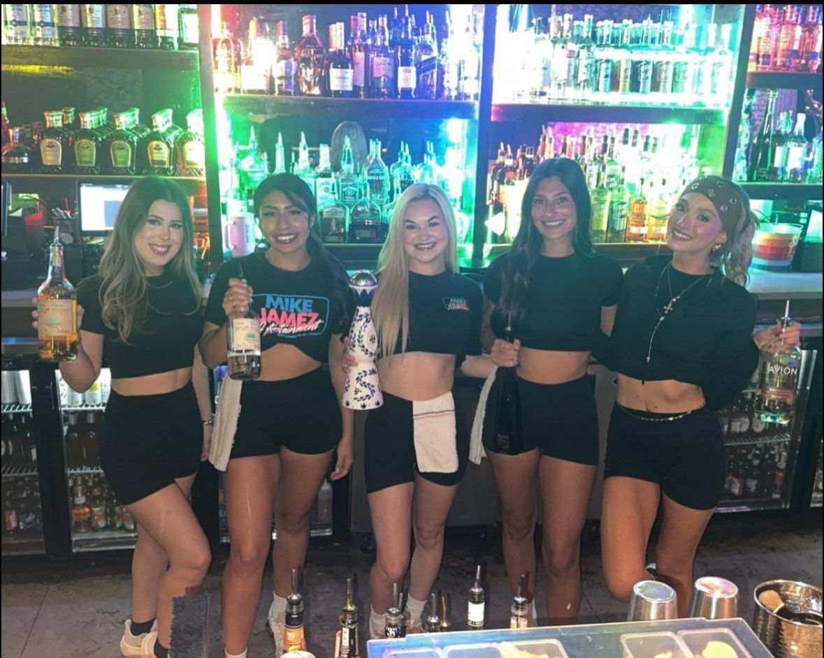 Five people posing with drinks behind a brightly lit bar, smiling in black outfits.