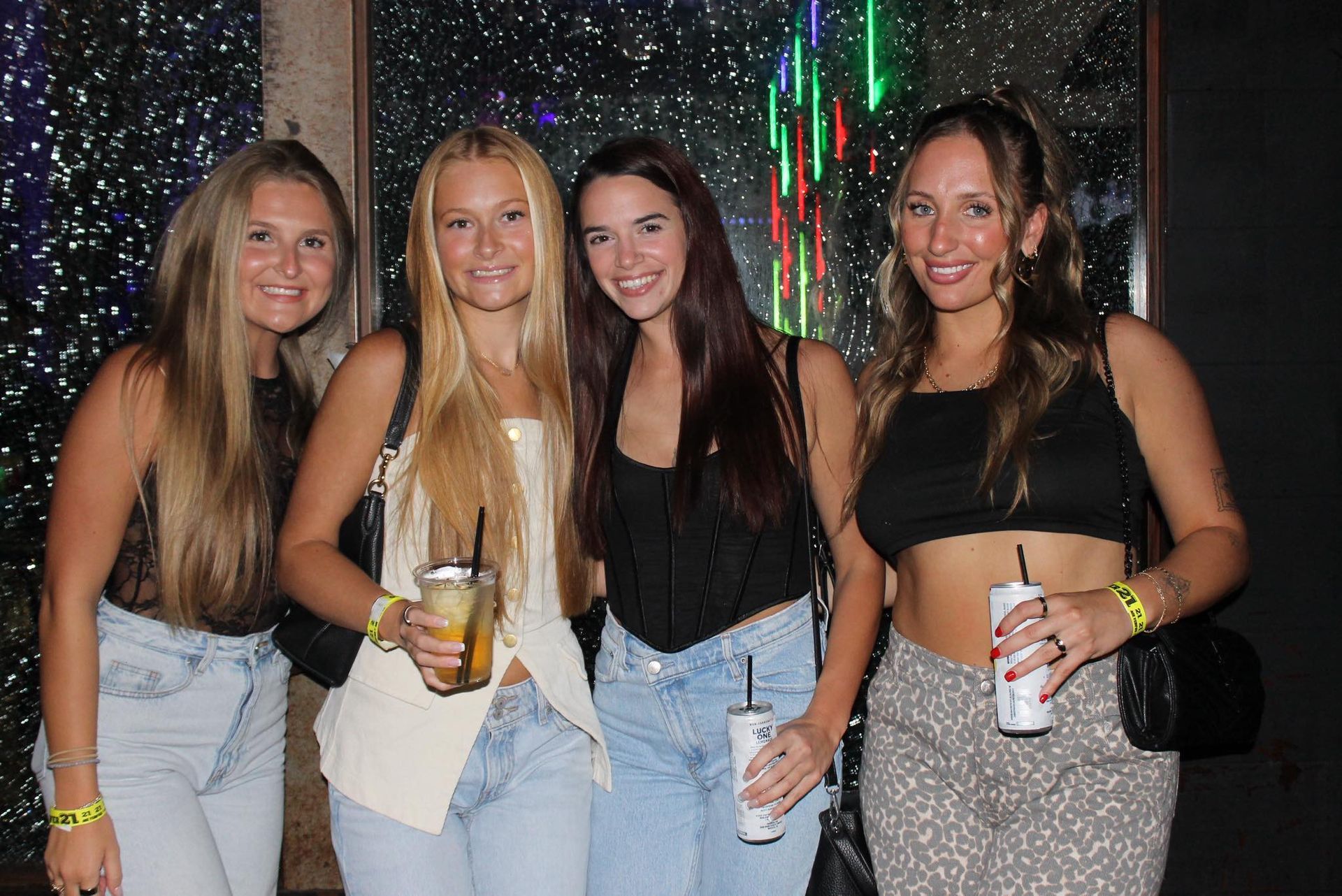 Four women posing at a party, holding drinks, in front of a sparkling backdrop.