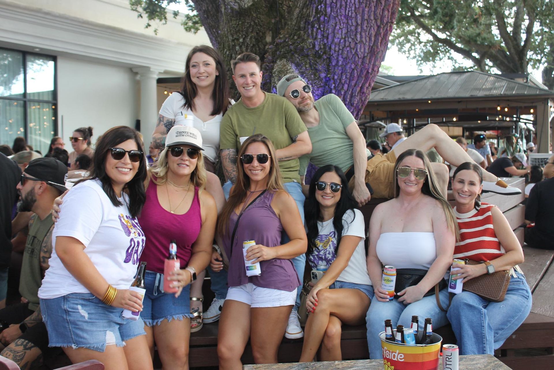 Group of friends posing outdoors at a patio table, smiling and holding drinks.