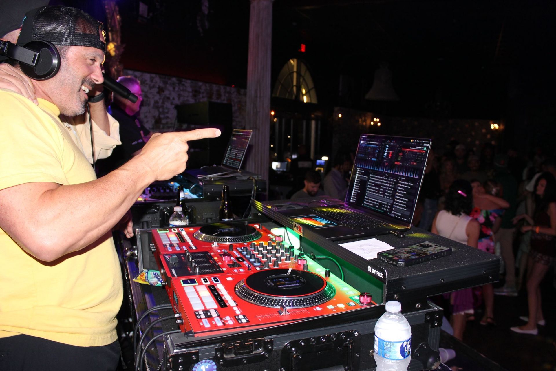 DJ in yellow shirt performing at a turntable setup in a dark club, pointing toward the crowd