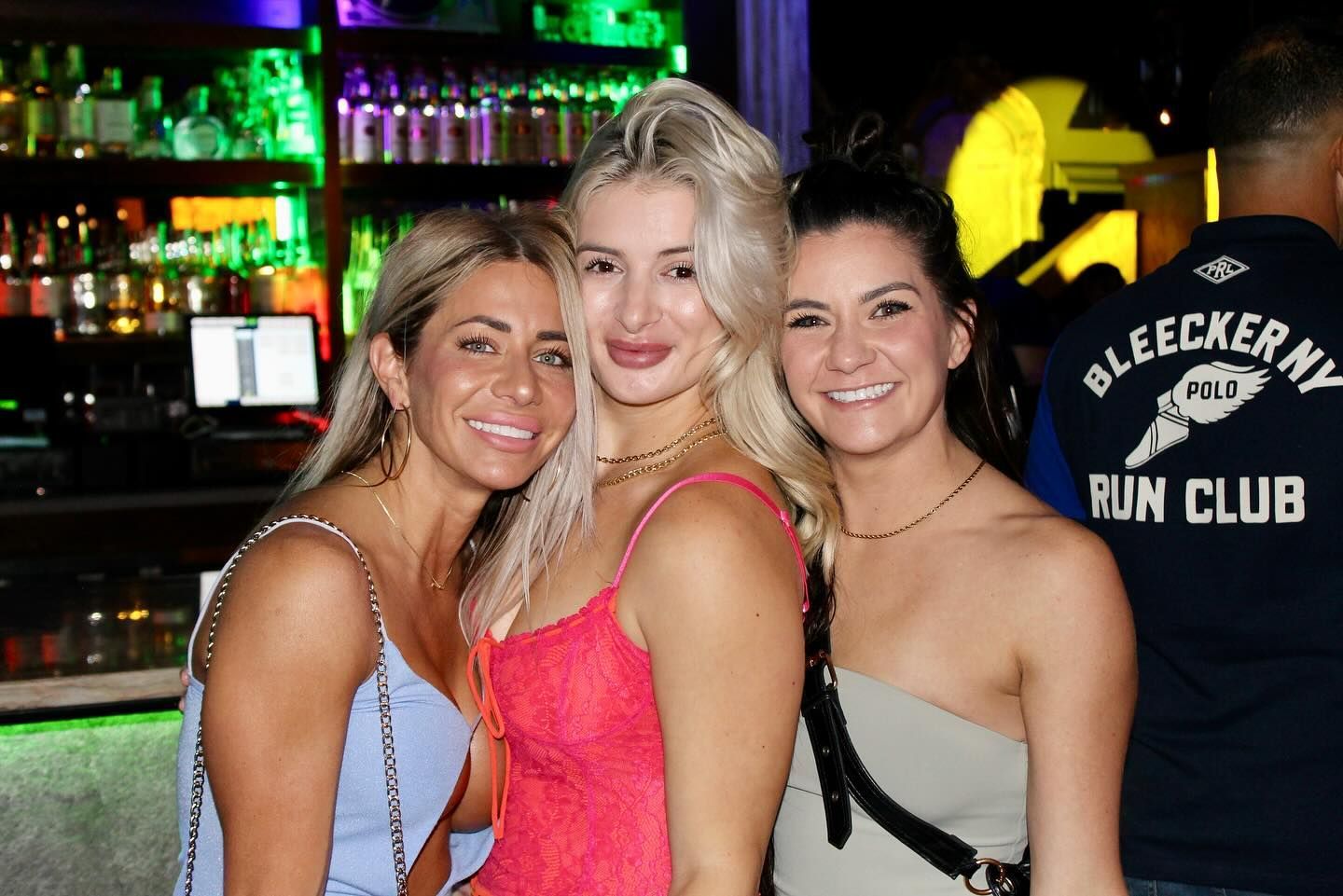 Three women smiling at a nightclub bar, with bottles behind them and a Bleecker Street Run Club jacket visible.