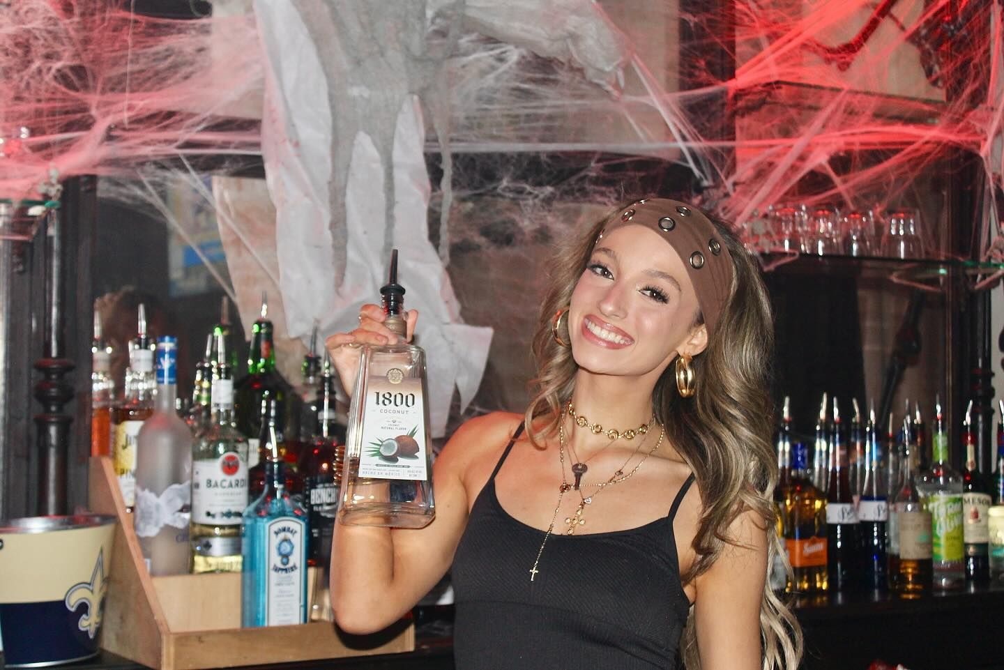 Woman smiling behind a bar, holding up a bottle amid shelves of liquor and red streamers.