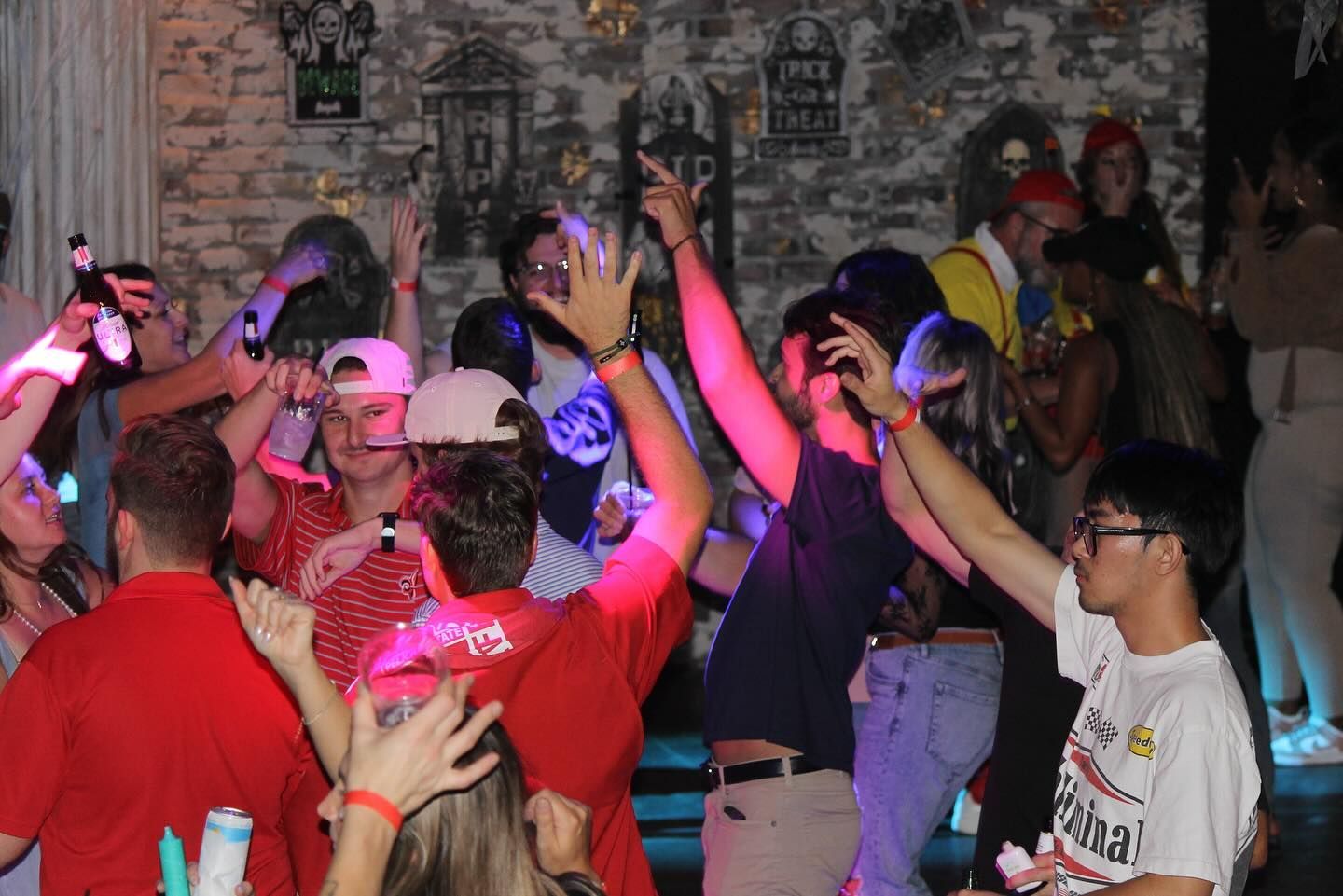 Crowd dancing at a party under pink lights, with people raising their hands indoors.