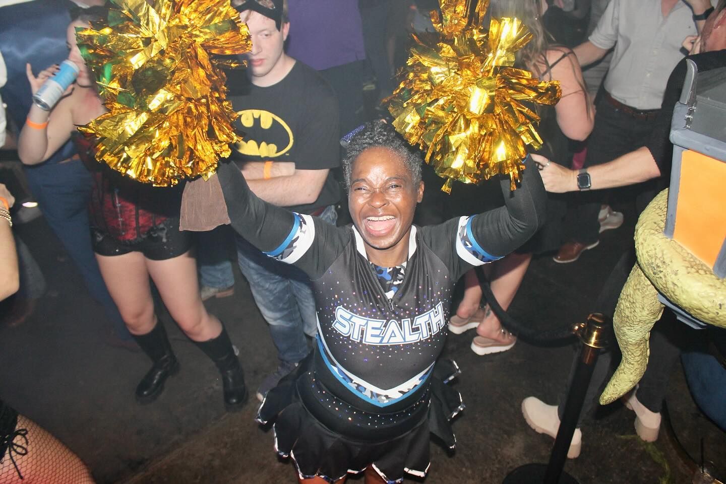 Cheering dancer in black outfit with gold pom-poms at a lively indoor party