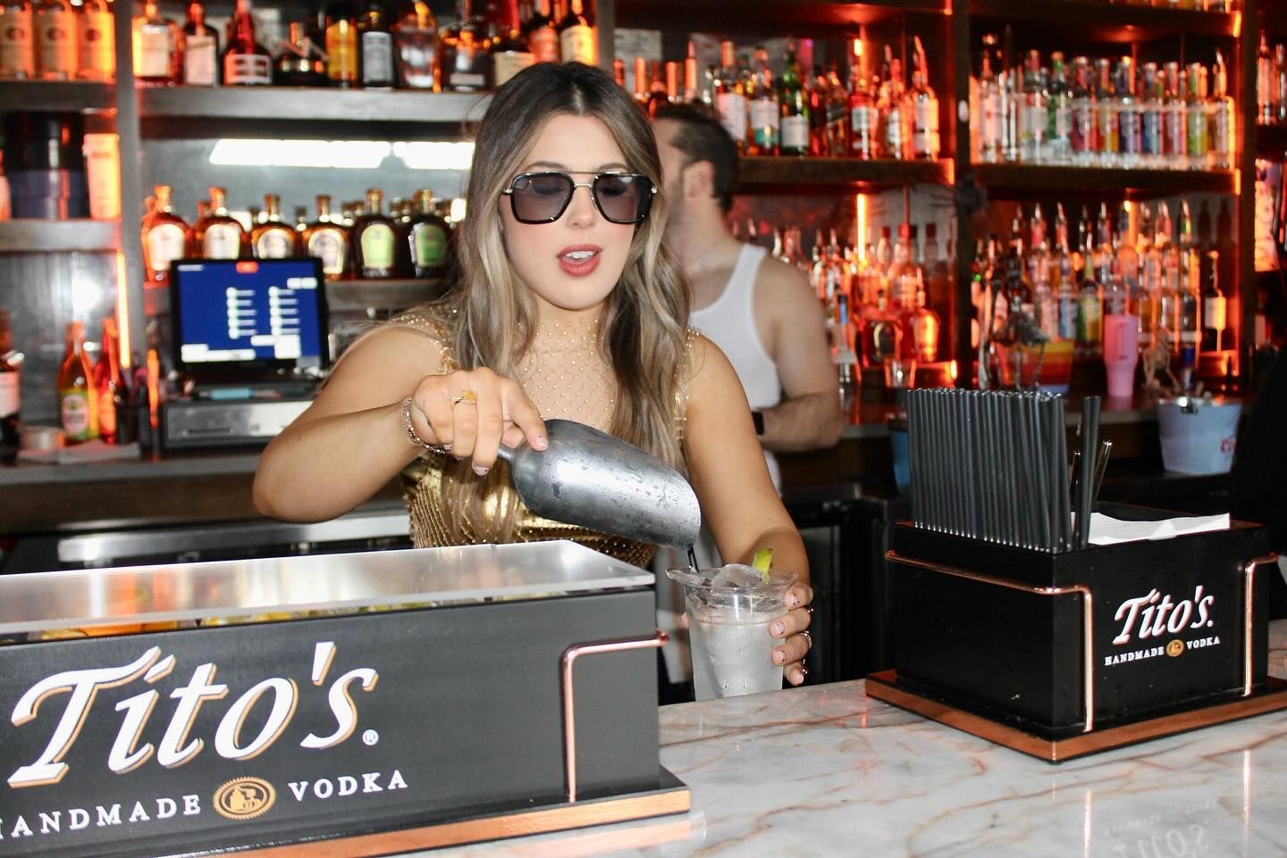 Bartender pouring a drink at a Tito's Vodka bar, with shelves of liquor bottles behind her.