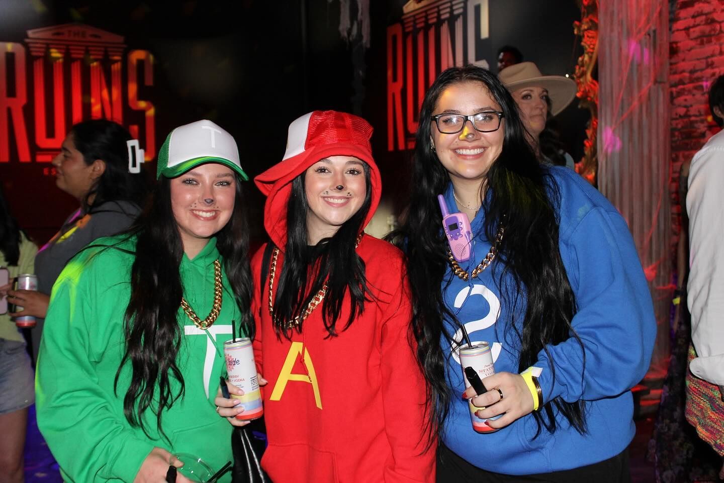 Three people in colorful costumes posing at an indoor event, smiling and holding drinks