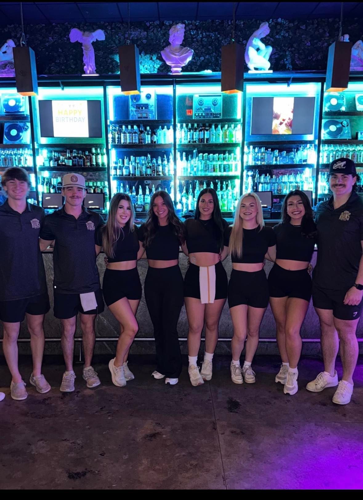 Group posing in front of a glowing bar shelf with blue and purple lights, wearing black outfits