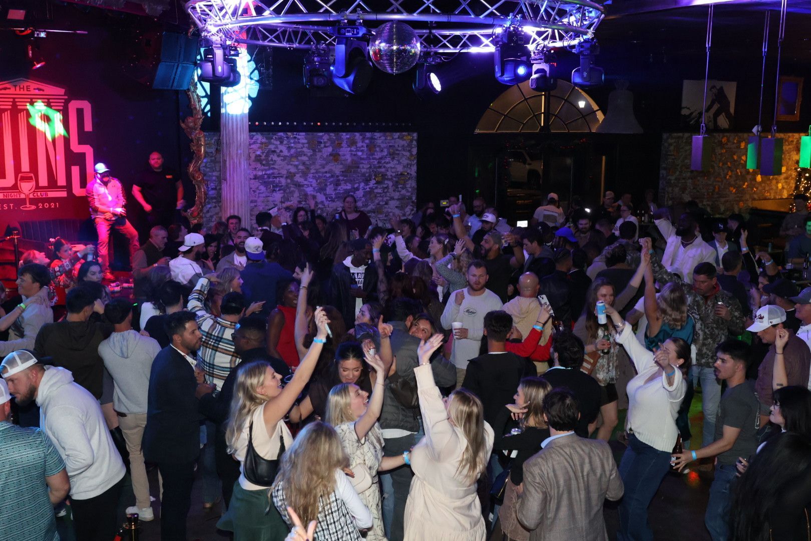 Crowded nightclub dance floor with people raising their hands under colorful stage lights