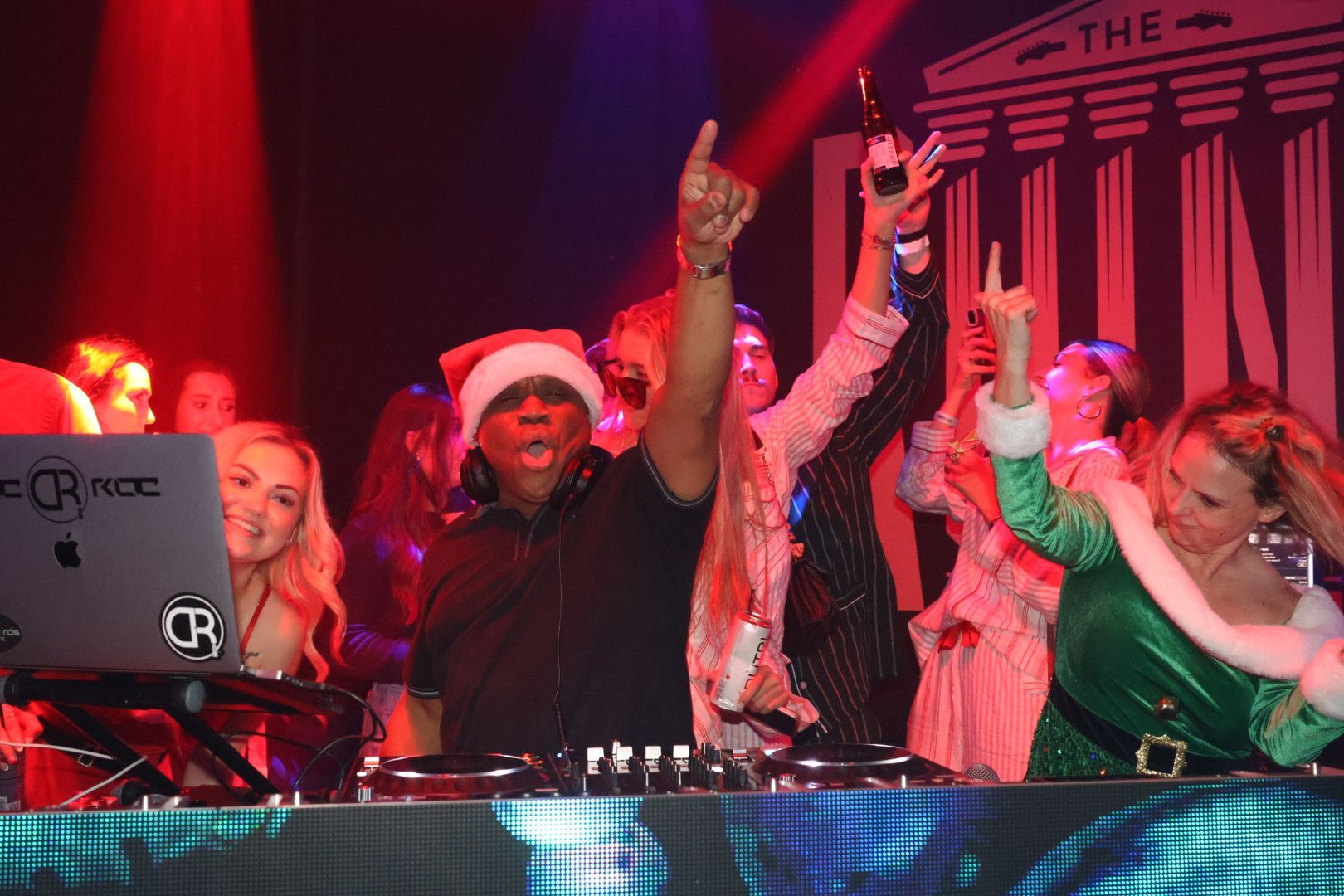 DJs in Santa hats dancing at a red-lit holiday club party with raised hands behind a DJ booth