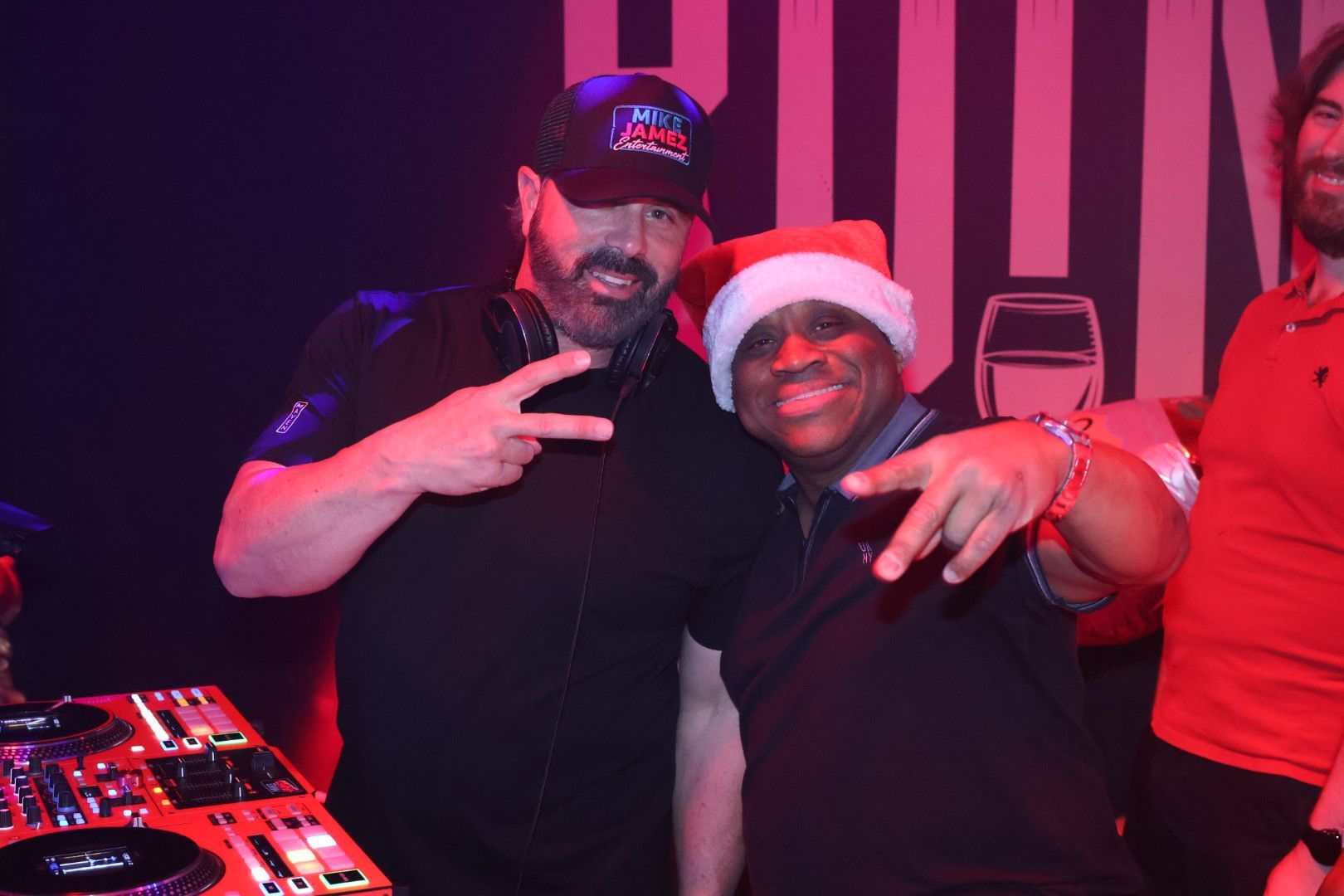 Two people smiling and posing with peace signs at a nightclub DJ booth, lit by pink and purple lights