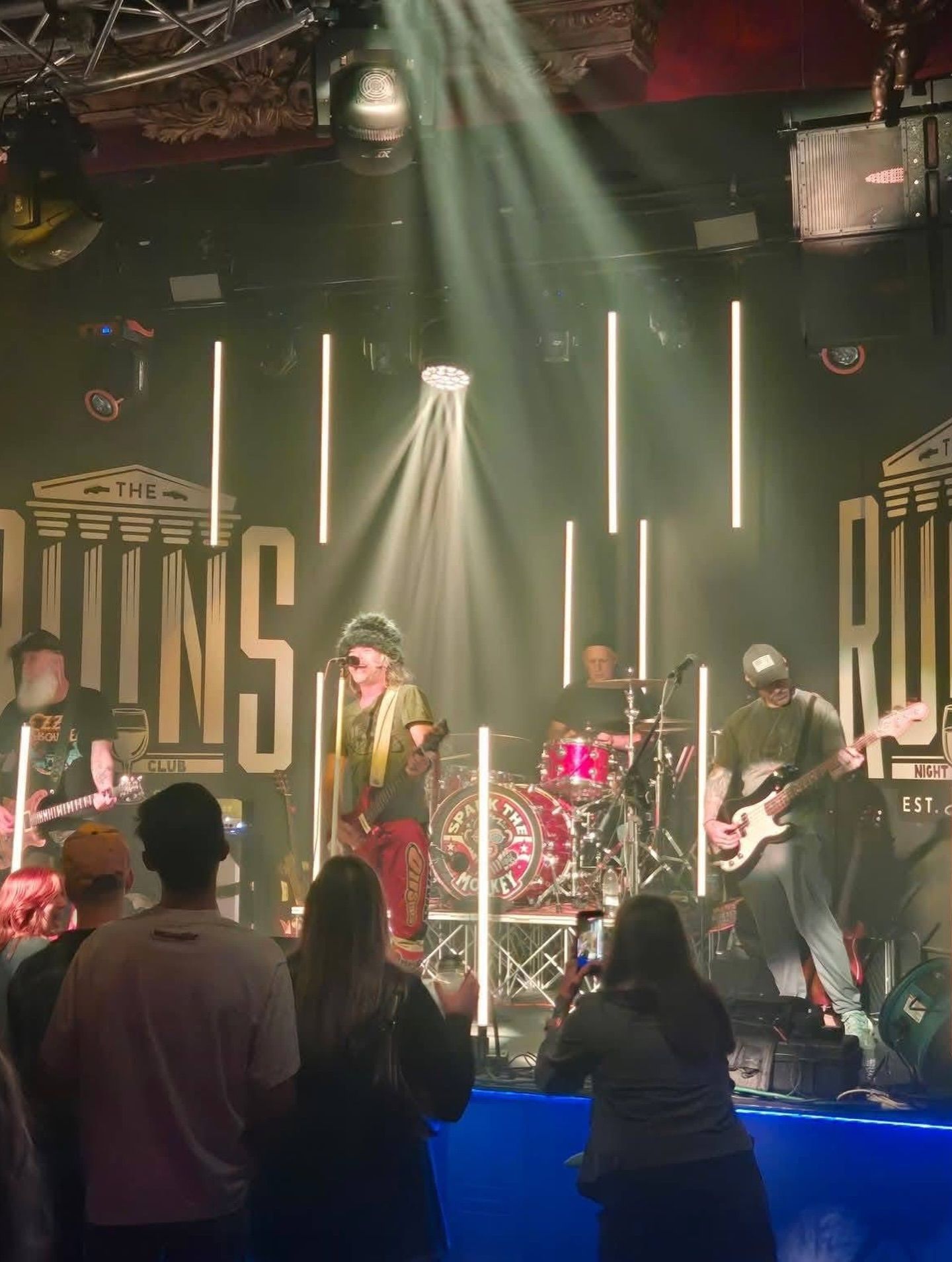 Live band performing on stage under green spotlights, with singer, guitarist, and drummer visible.