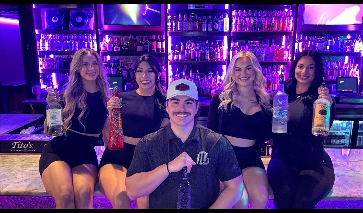Five friends seated at a neon-lit bar, smiling and holding colorful drinks together