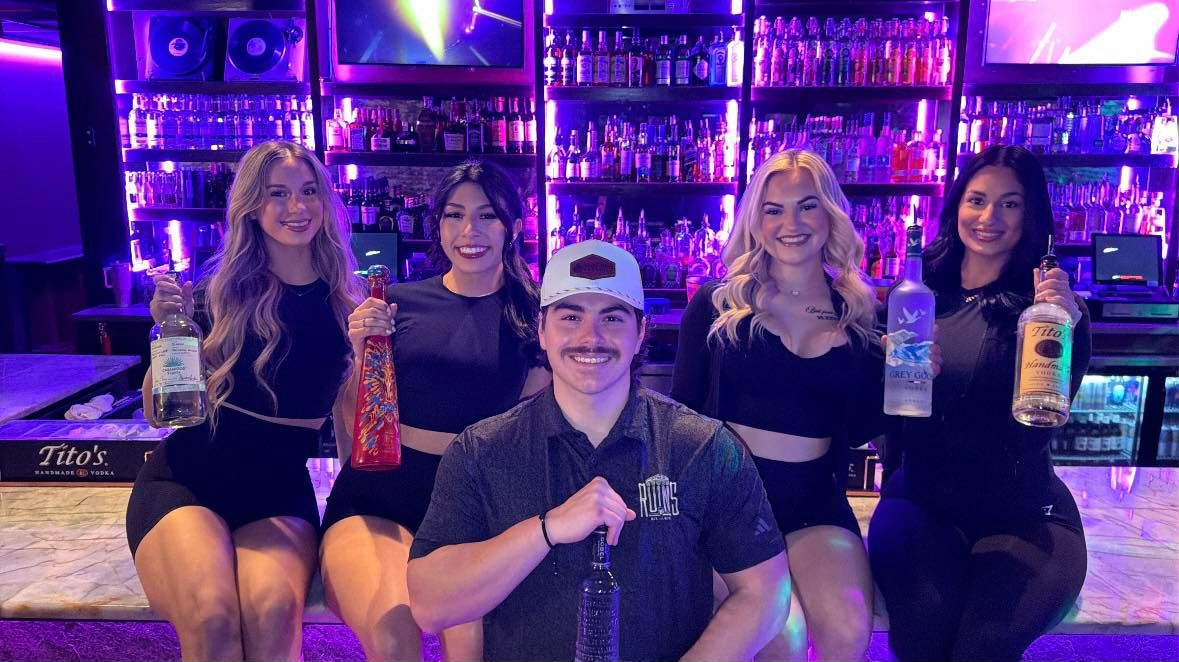 Five people posing at a neon-lit bar, holding drinks and smiling.