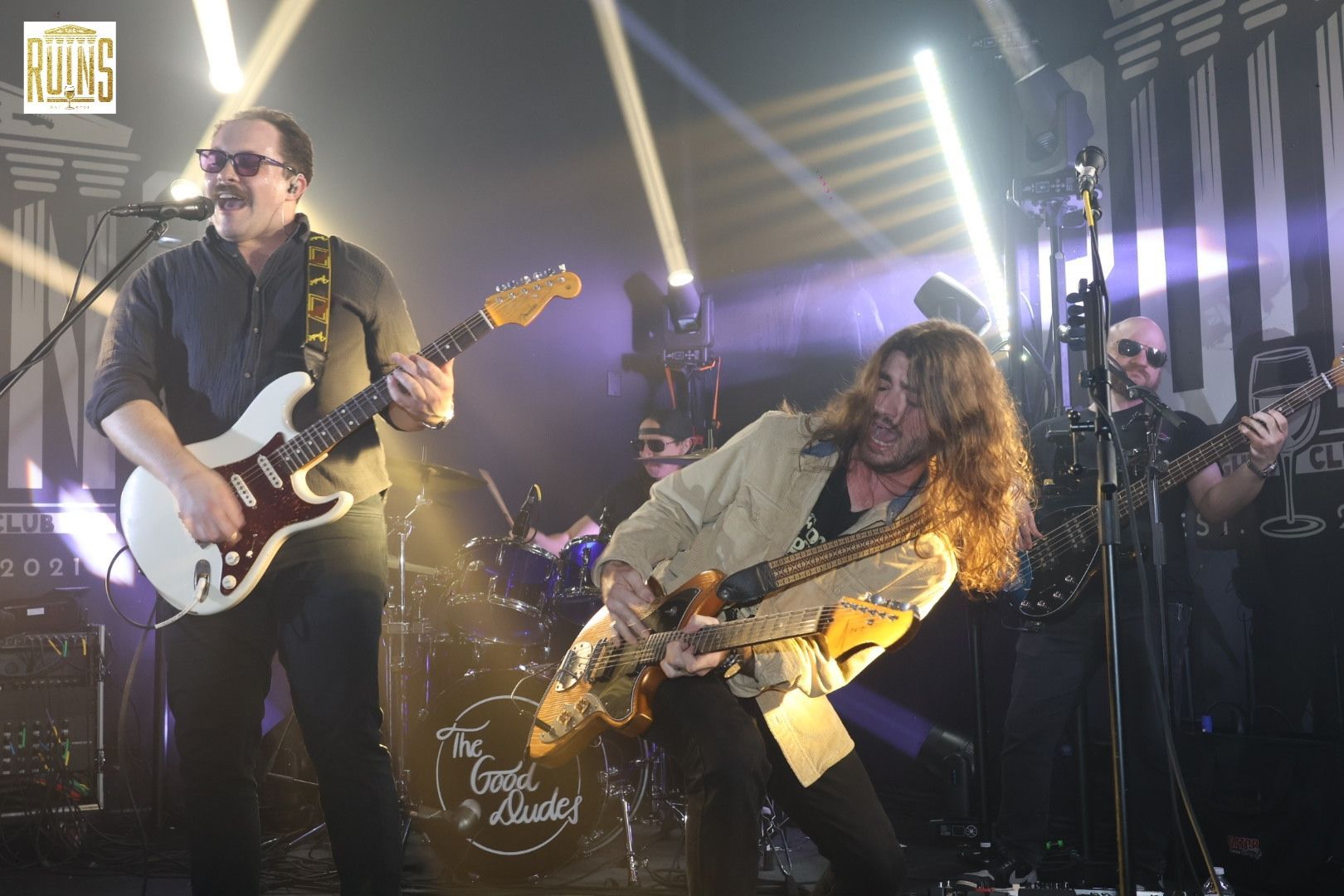 Musicians playing electric guitars on a brightly lit stage, with one leaning forward in an energetic performance.