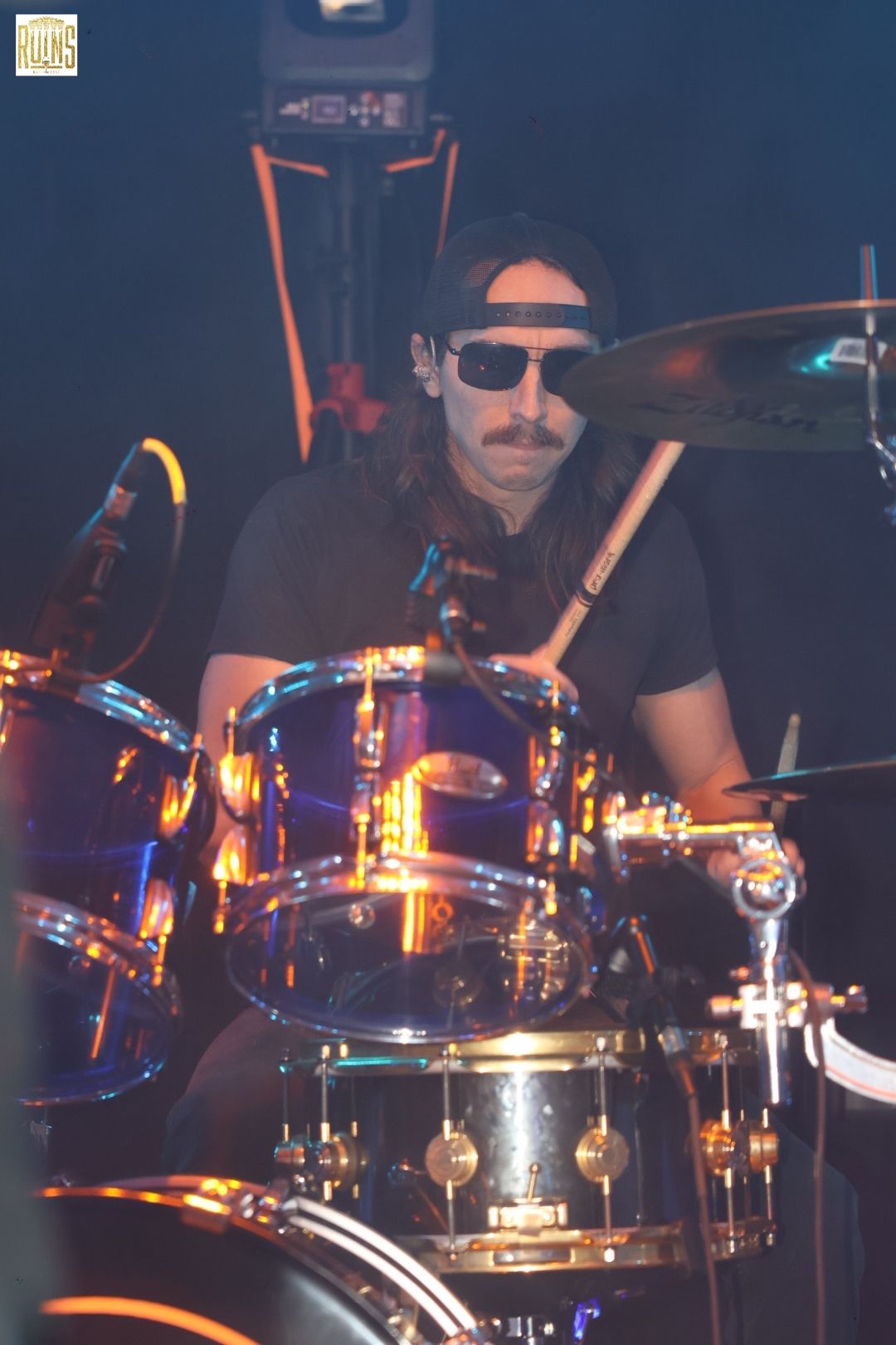 Drummer wearing sunglasses behind a shiny drum kit on a dimly lit stage