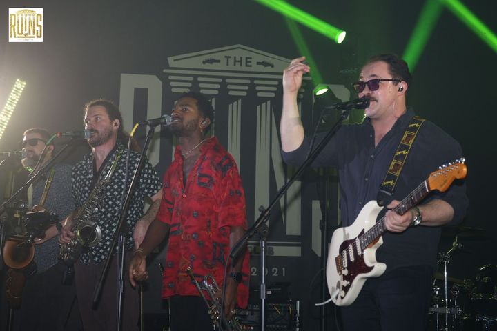 Live band onstage with saxophone, vocals, and electric guitar under green lights