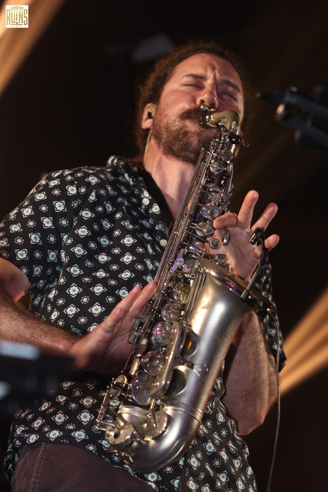 Musician playing a saxophone onstage, eyes closed, wearing a patterned shirt under warm stage lights