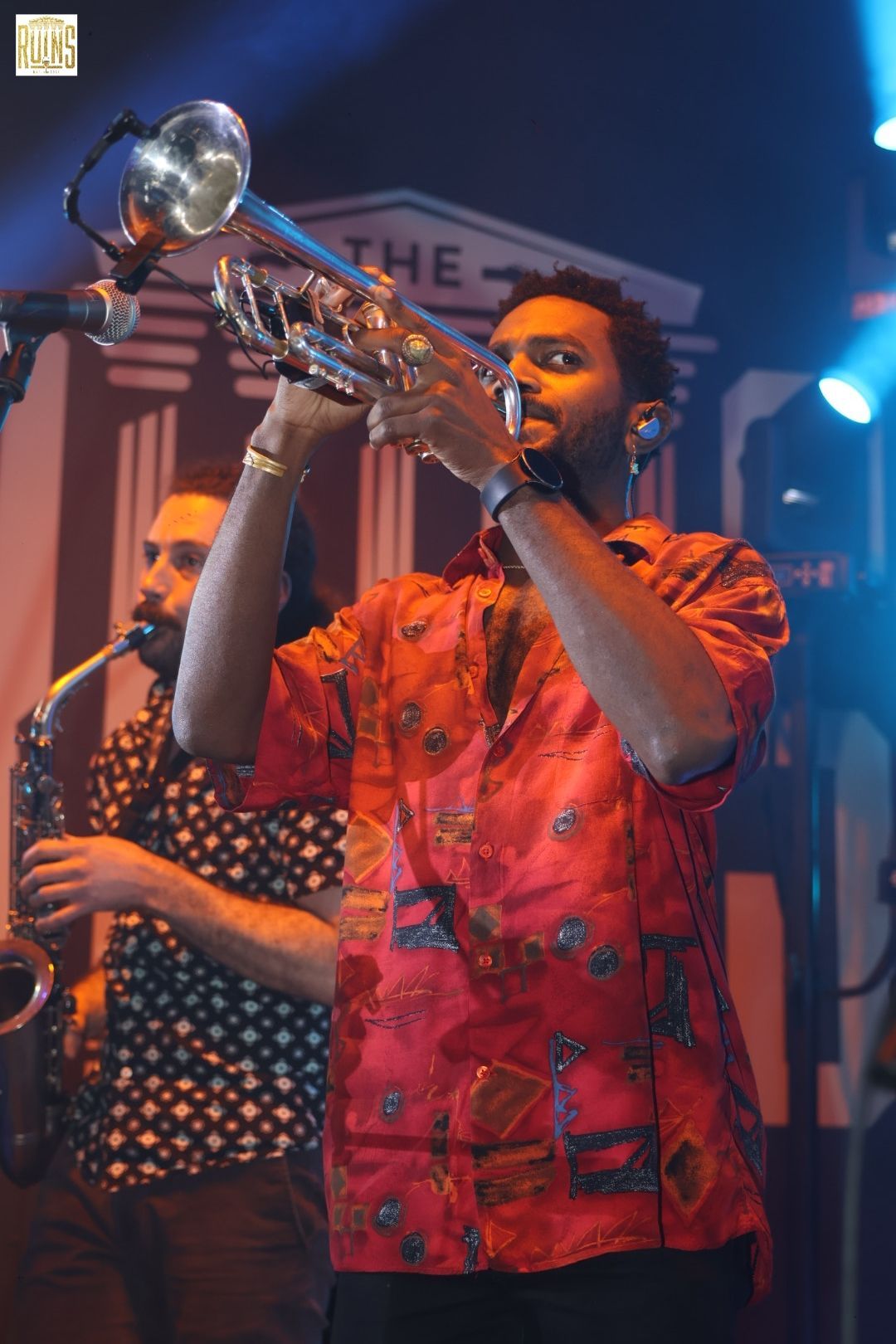 Musician in a red shirt playing trumpet onstage under blue and orange lights