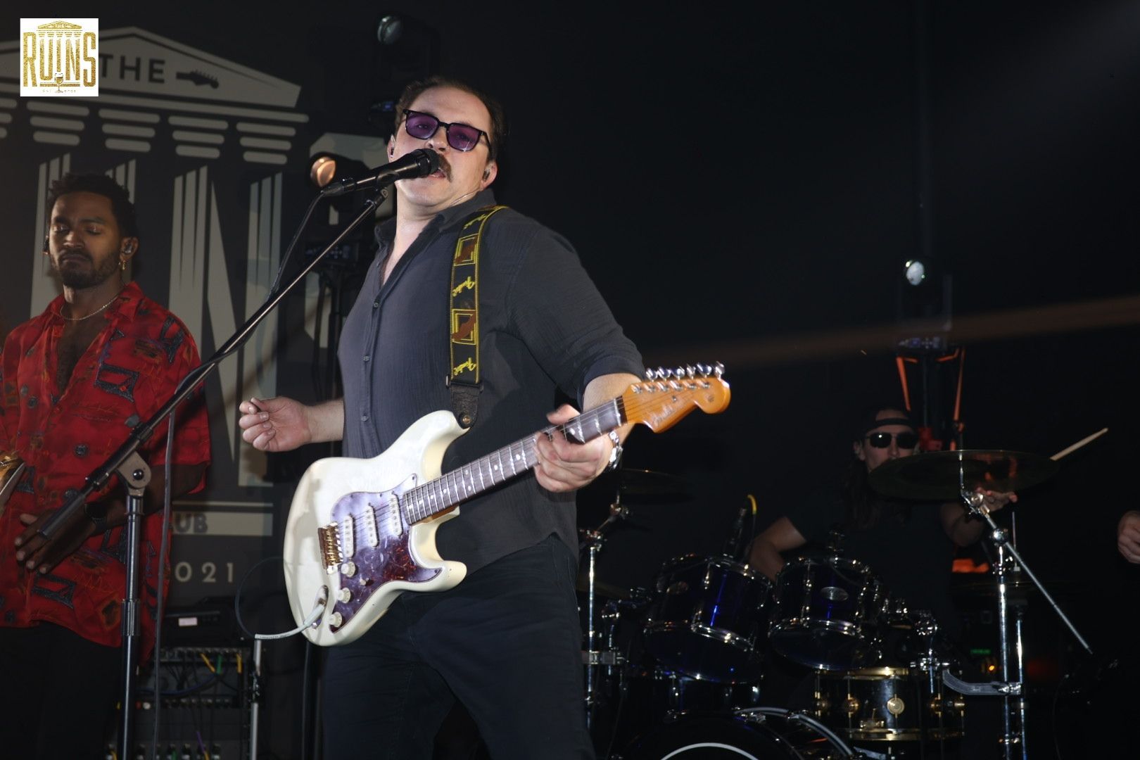 Singer playing white electric guitar on stage with drummer in the background, under low club lighting