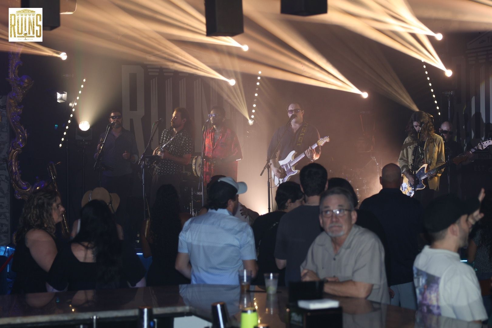 Live band performing on a dimly lit stage with bright spotlights, viewed from a crowded audience.