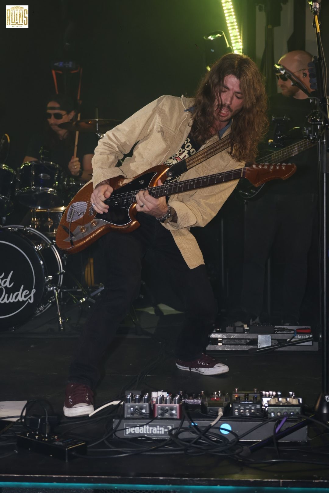 Guitarist in beige shirt leaning back on stage, playing electric guitar amid pedal board and dark concert lighting.