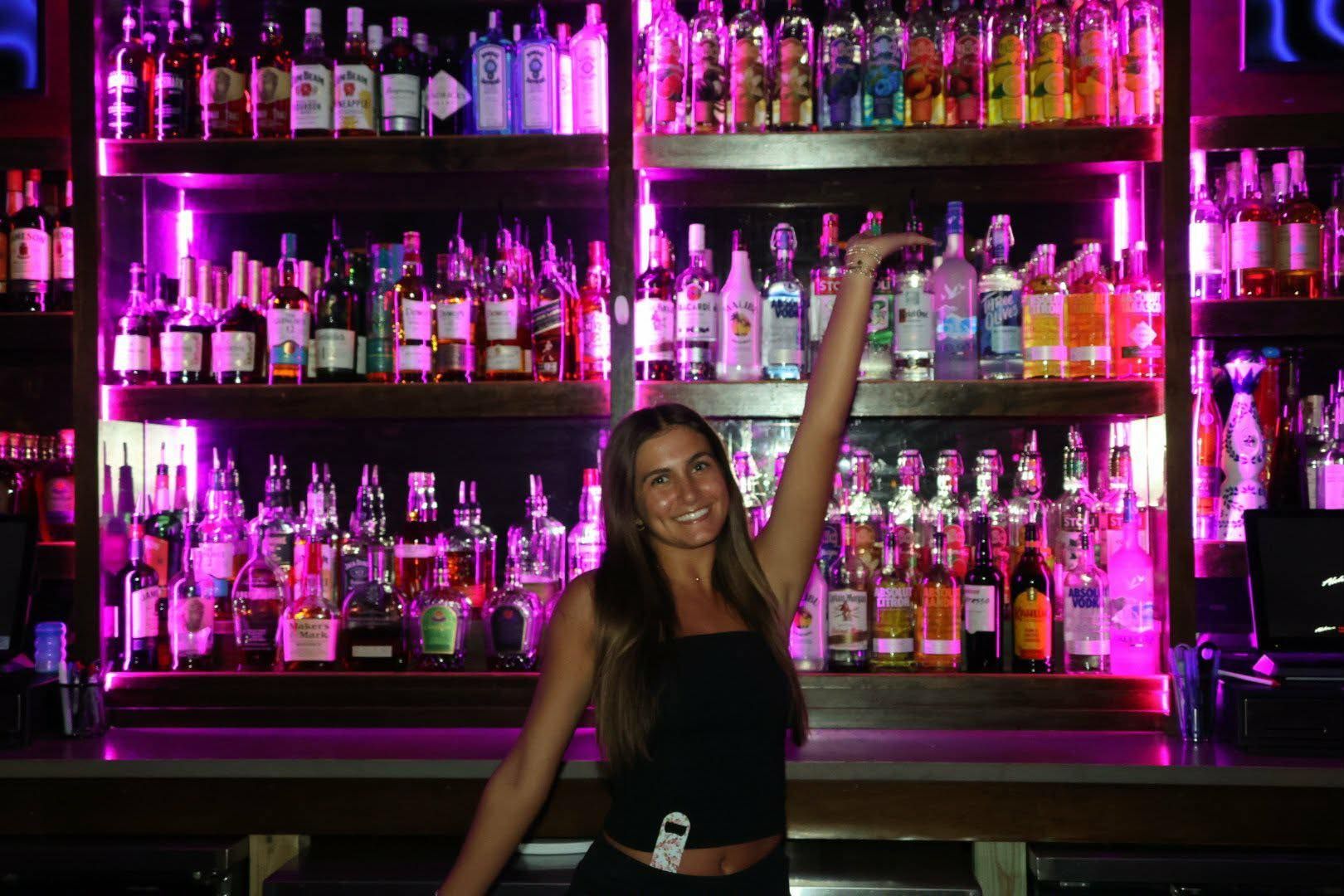 Woman posing in a neon-lit bar in front of shelves filled with colorful bottles