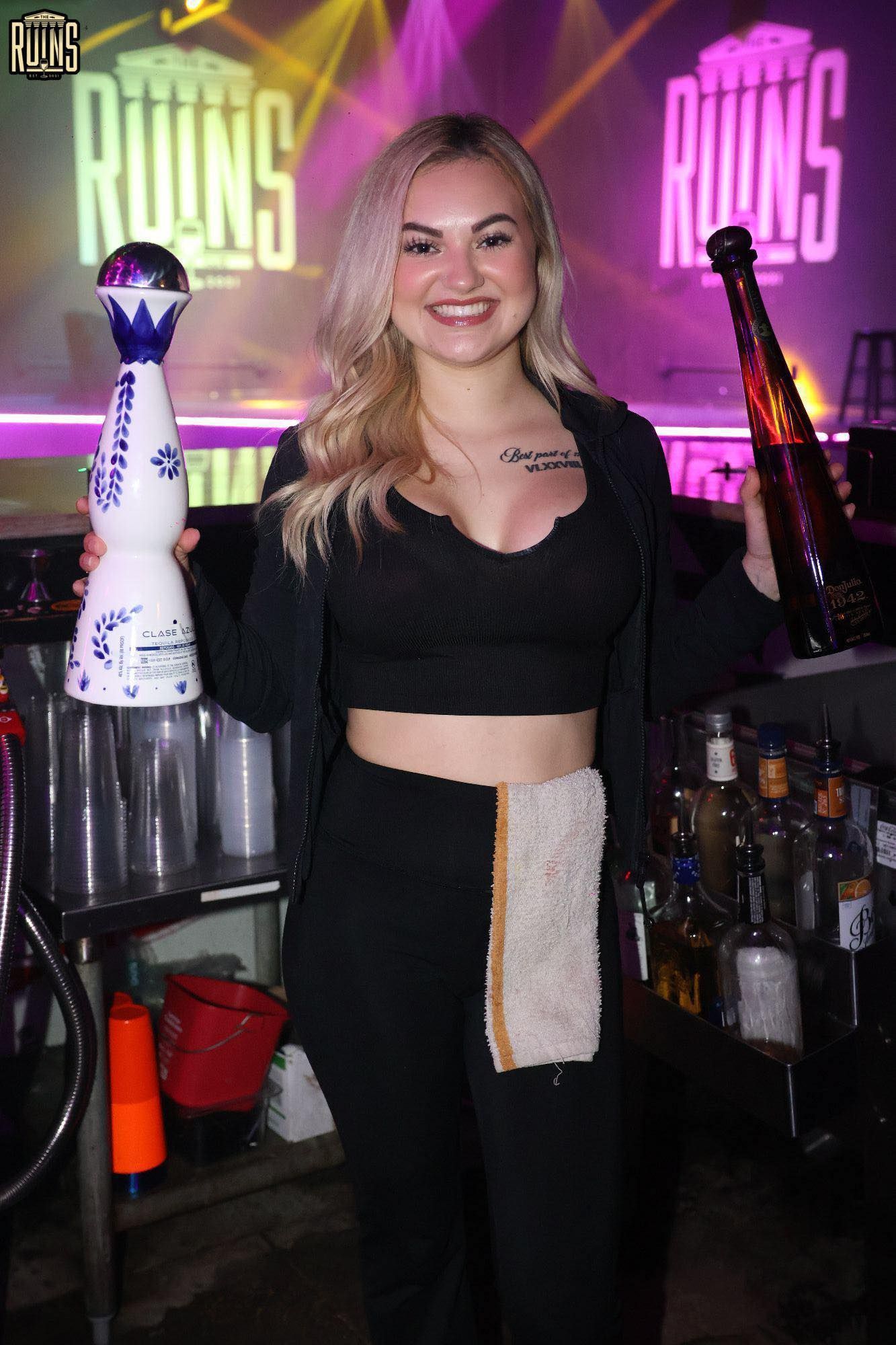 Woman in black outfit posing behind a bar with purple neon lights and liquor bottles.