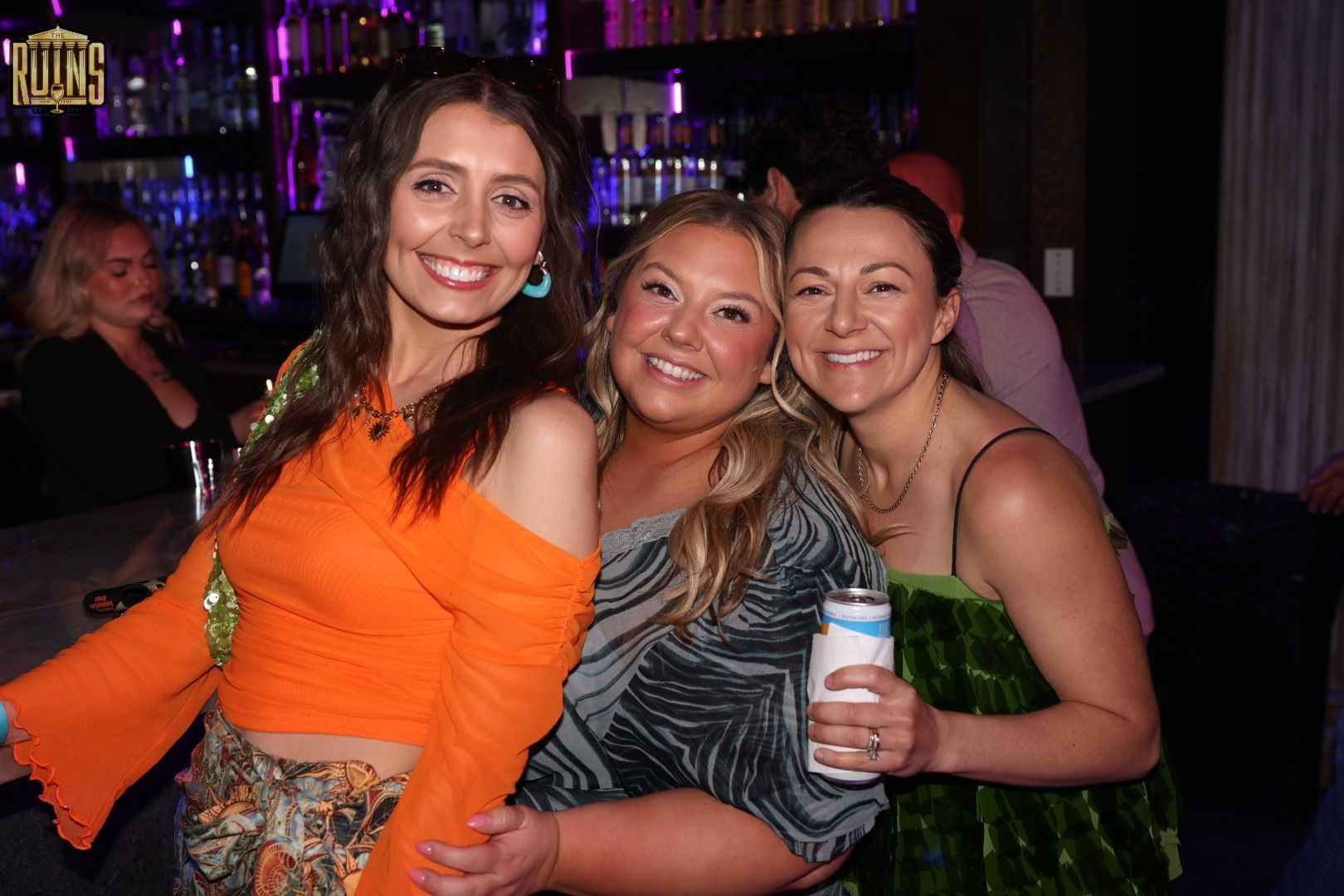 Three women smiling and posing together at a dimly lit party, with drinks in hand.
