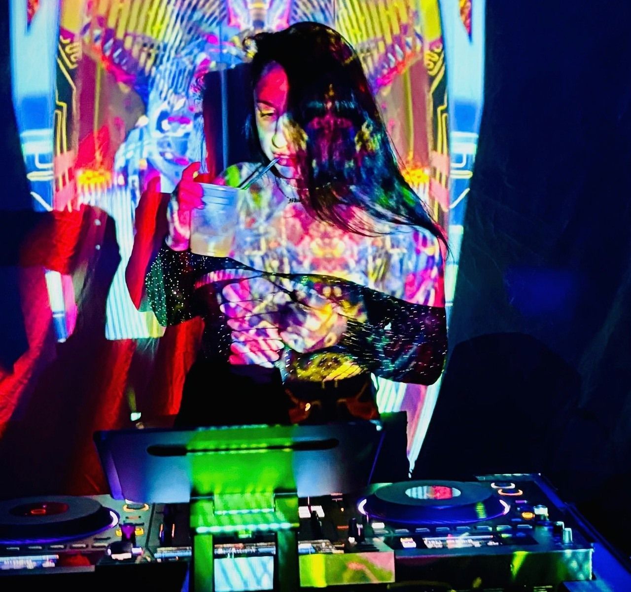 DJ performing at a neon-lit booth, holding a drink amid colorful projected lights and turntables