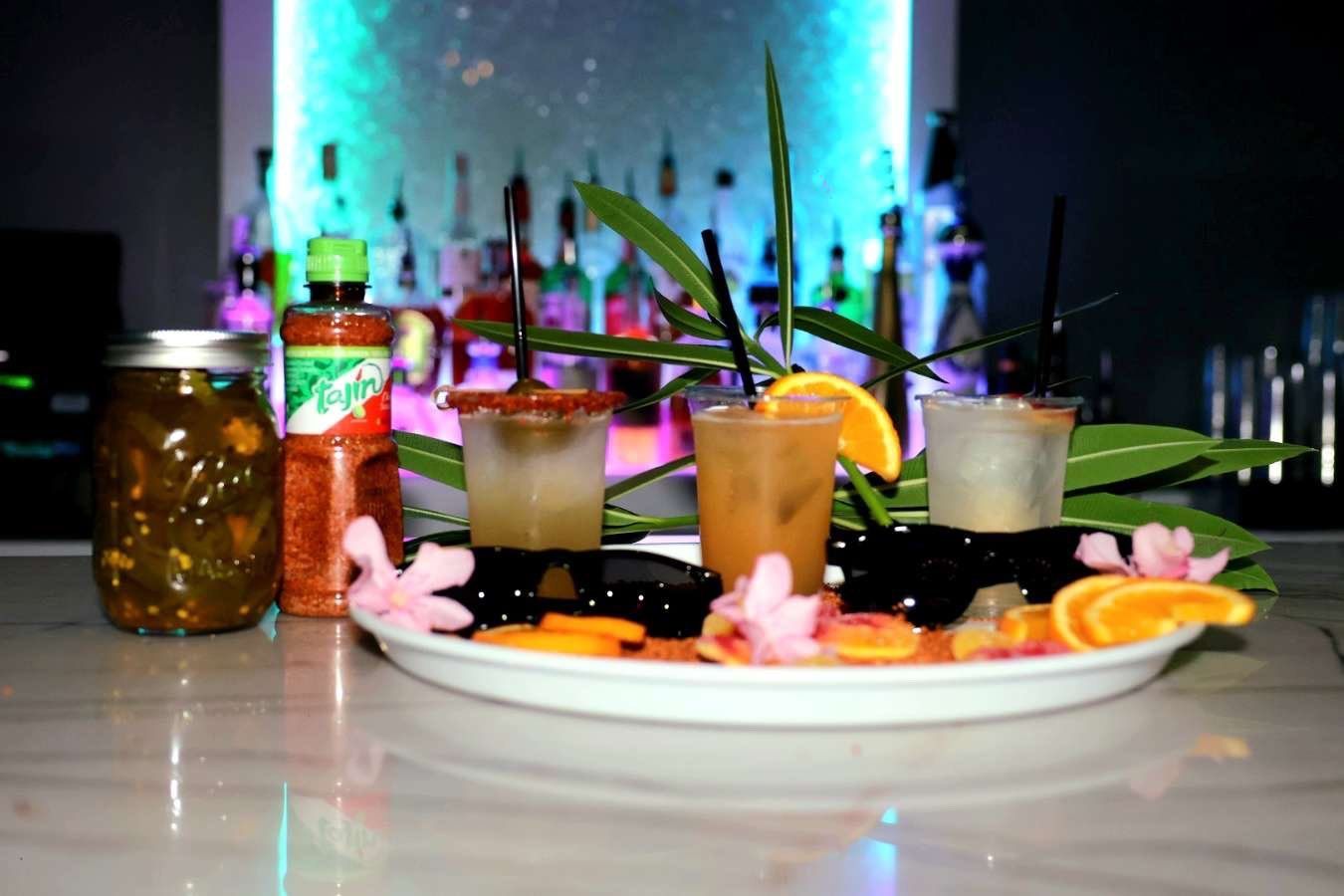Cocktail tray with colorful drinks, citrus garnishes, and flowers on a bar counter at a dimly lit lounge