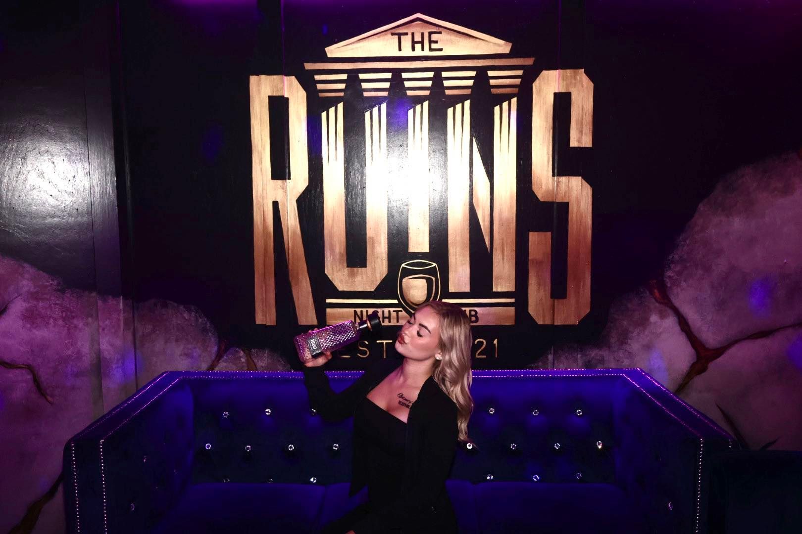 Person posing in front of a large “The Ruins” sign with purple lighting and a black couch