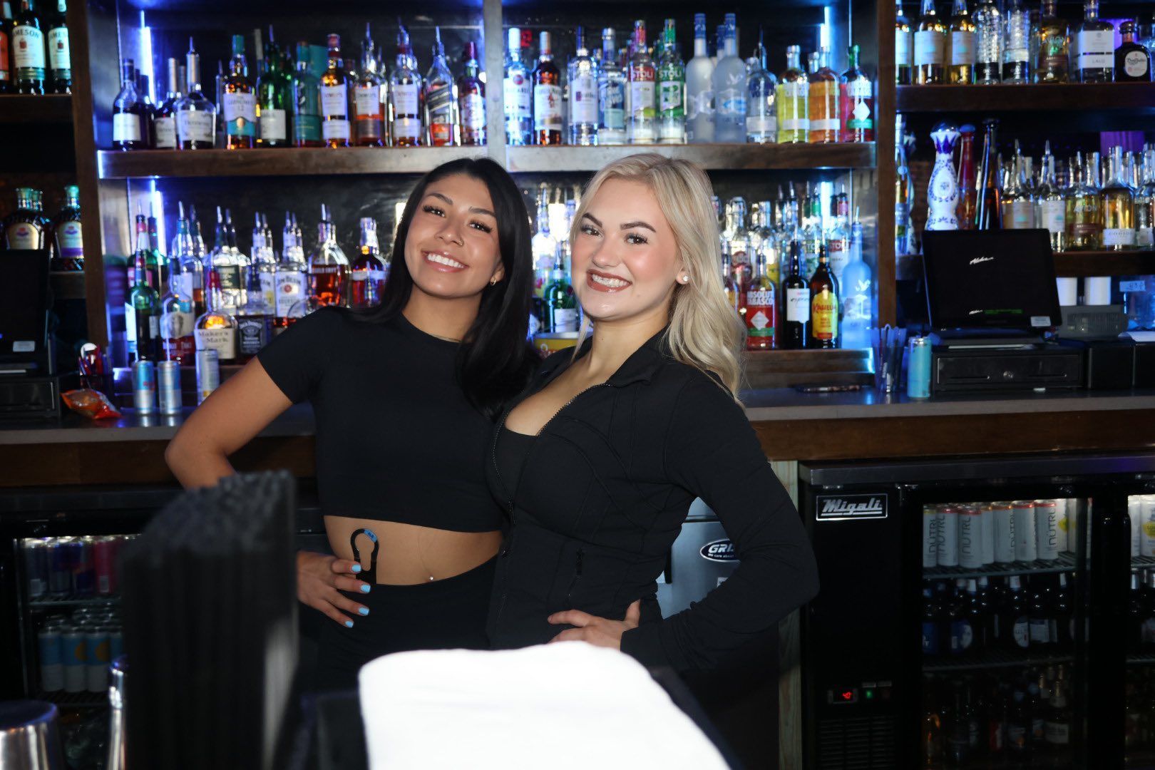 Two women posing and smiling in a bar, with shelves of liquor bottles behind them.
