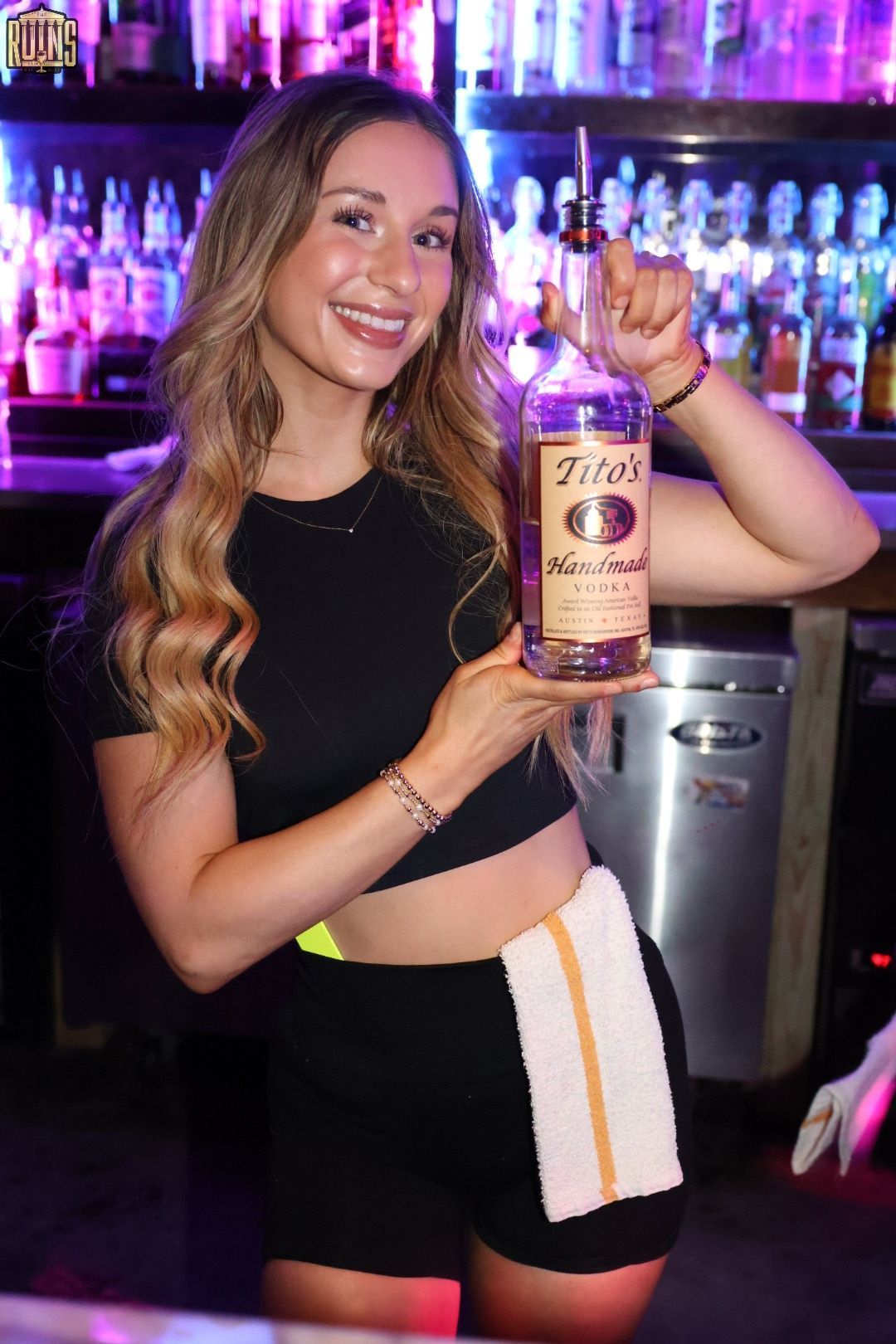 Woman in black top and shorts holding a bottle behind a bar with purple lighting, smiling at the camera